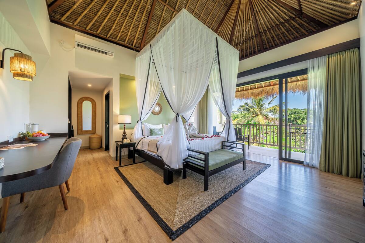 A bedroom with a bed, a dining table, a chair, a nightstand, a lamp, and a view of the outdoors.