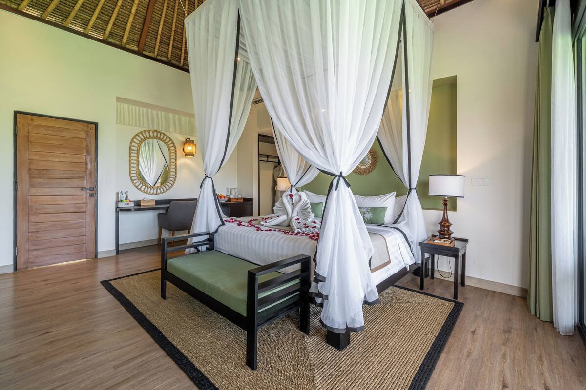 A bedroom with a four-poster bed, a bedside table, a chair, a desk, a mirror, and a door.
