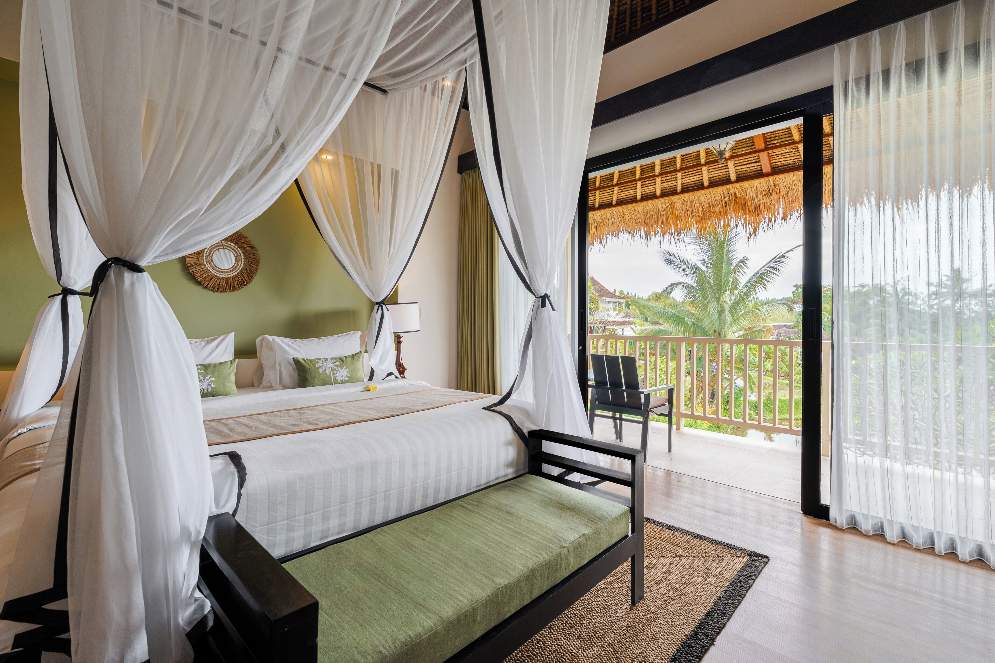 A bedroom with a bed, a bench, a chair, a window with a view of palm trees, and a balcony.