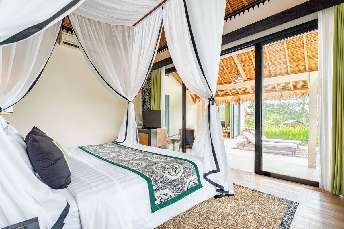 A four-poster bed with a patterned bedspread, a television on a stand, and a view of the outdoor patio area.