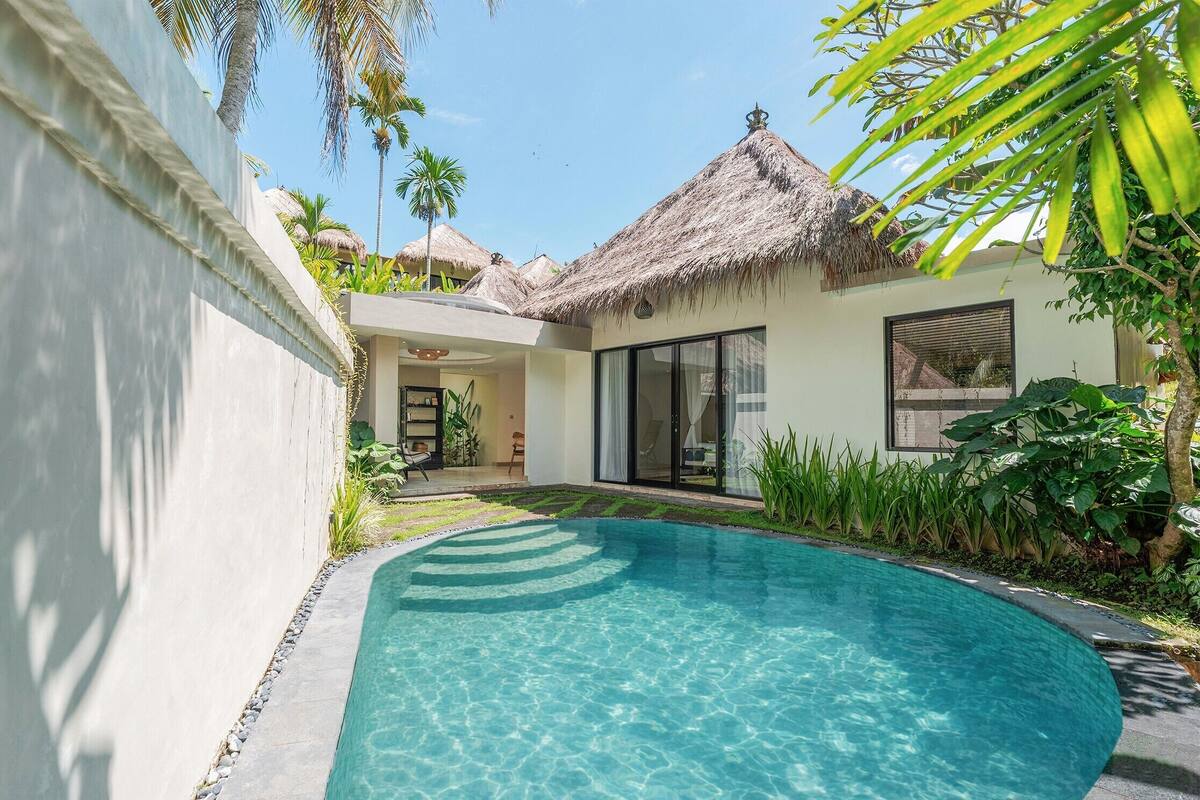 A pool with a curved edge, a thatched-roof structure, and a modern building with large windows.