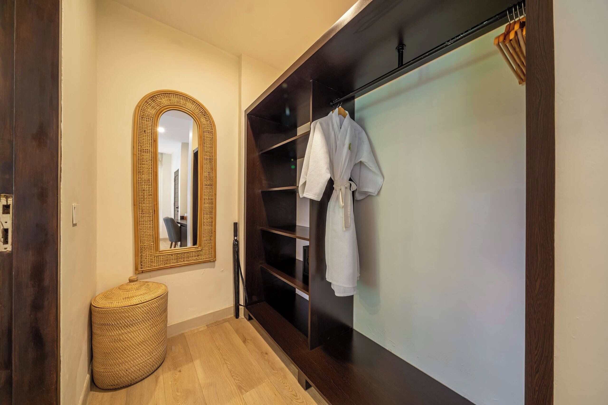 A hallway with a mirror, a wicker basket, and a dark wooden closet with clothes hanging inside.