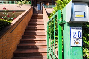 Eine Treppe mit grünem Metallgeländer, einem Schild mit der Aufschrift 'VIA DEL CASTRUCCIO 50' und einem Verteilerkasten mit 'LUCE SCALE'.