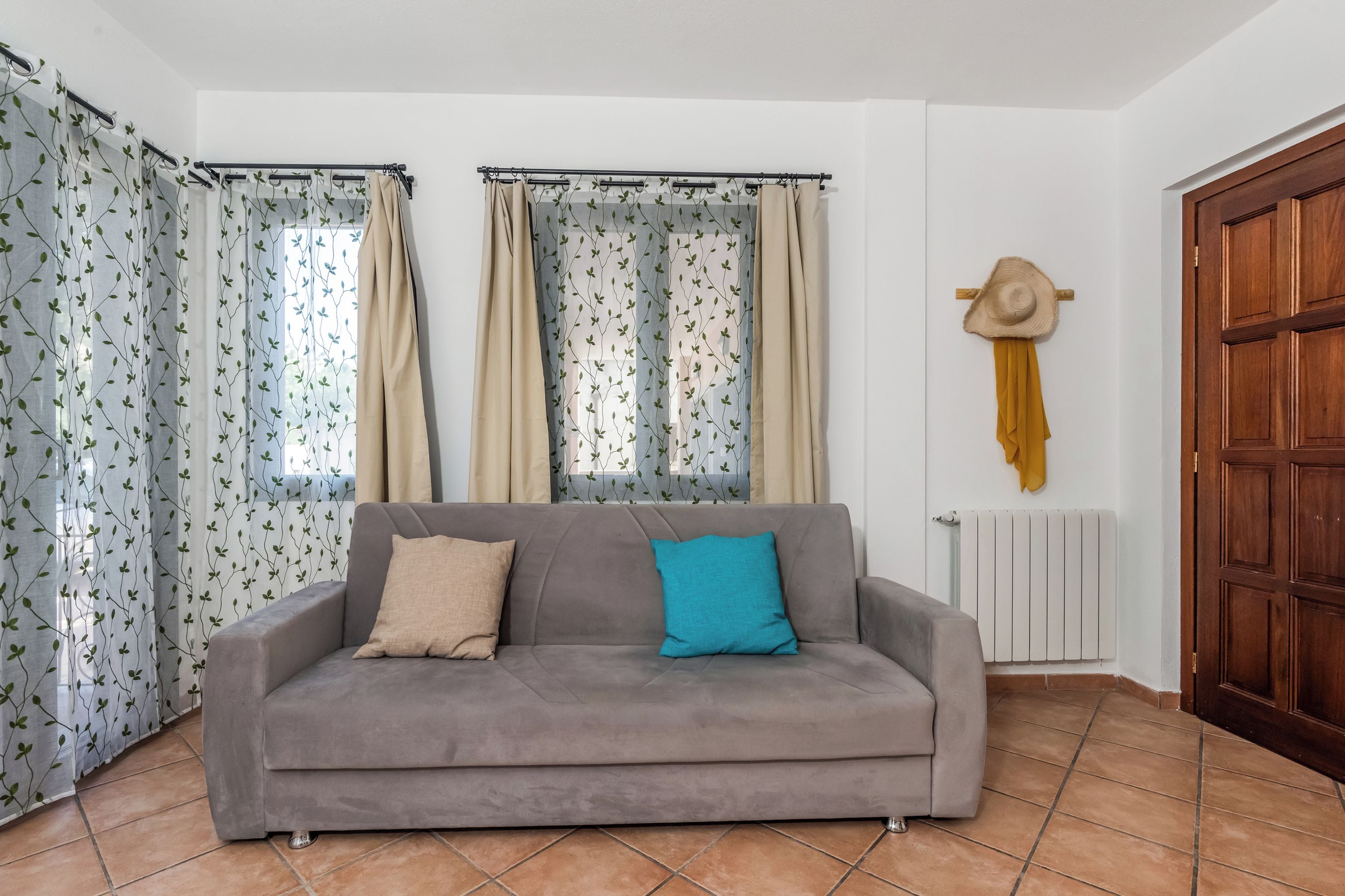 Un salon avec un canapé gris, des coussins beiges et bleus, une porte en bois et des rideaux à motifs.