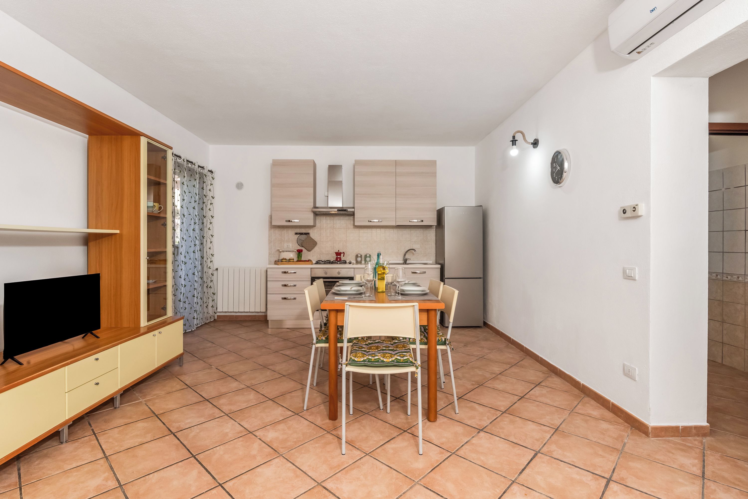 Una cocina compacta con área de comedor, un televisor sobre un soporte de madera y piso de baldosas.