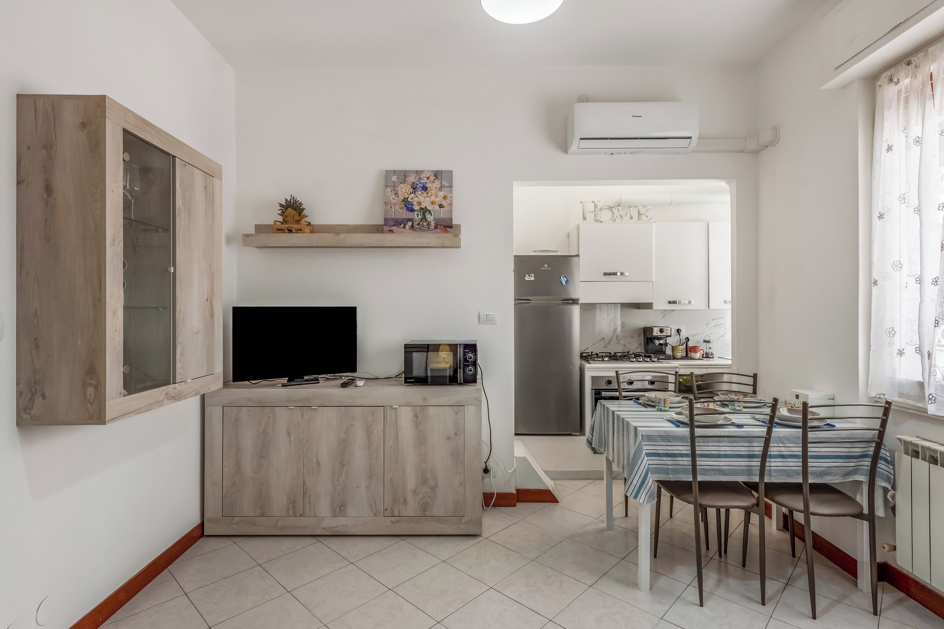 Cozinha compacta equipada com micro-ondas, fogão e frigorífico. Inclui mesa de jantar com cadeiras e uma televisão sobre um móvel de madeira.