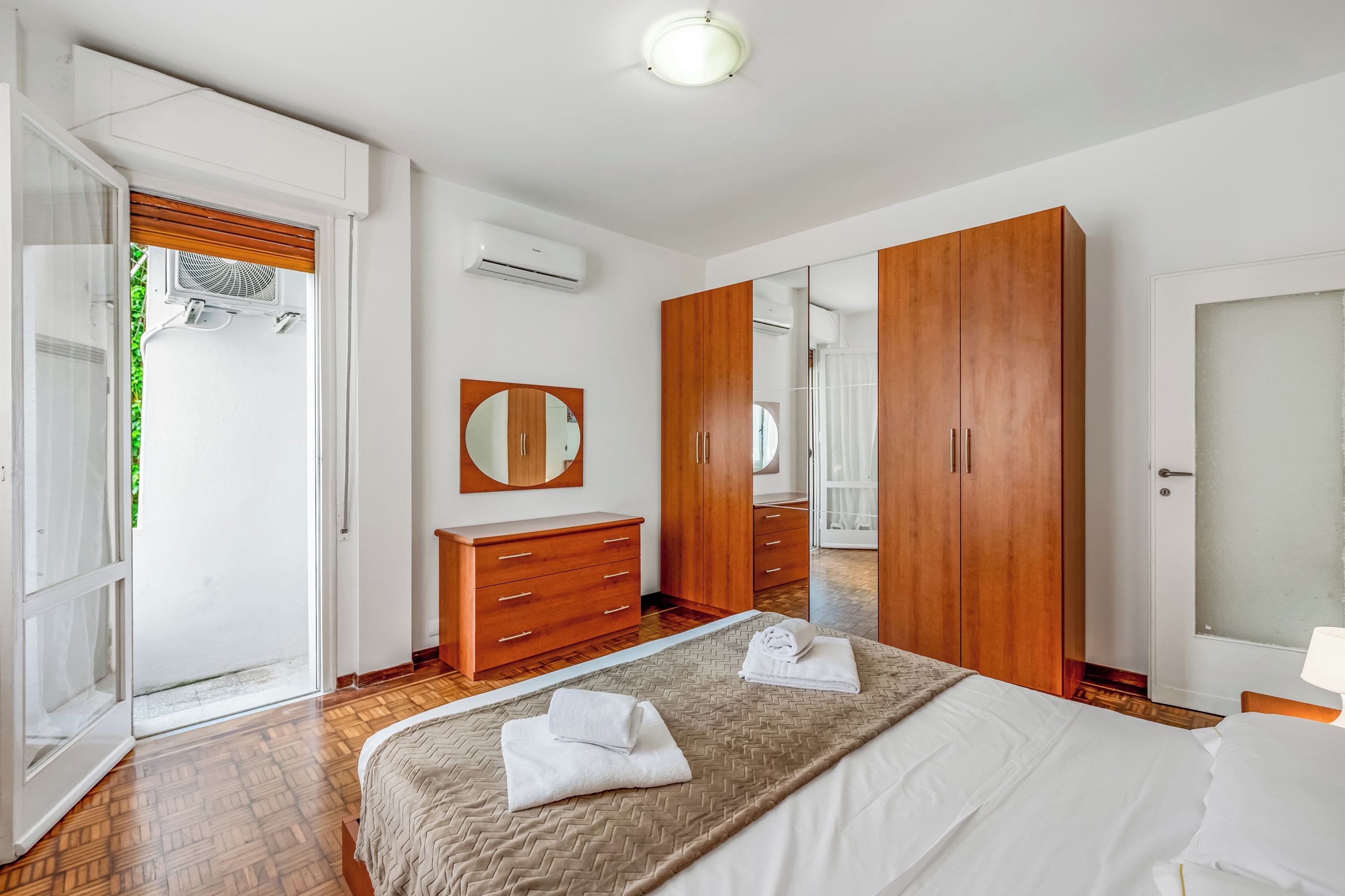 Quarto com cama, guarda-roupa de madeira, espelho e porta para uma varanda.