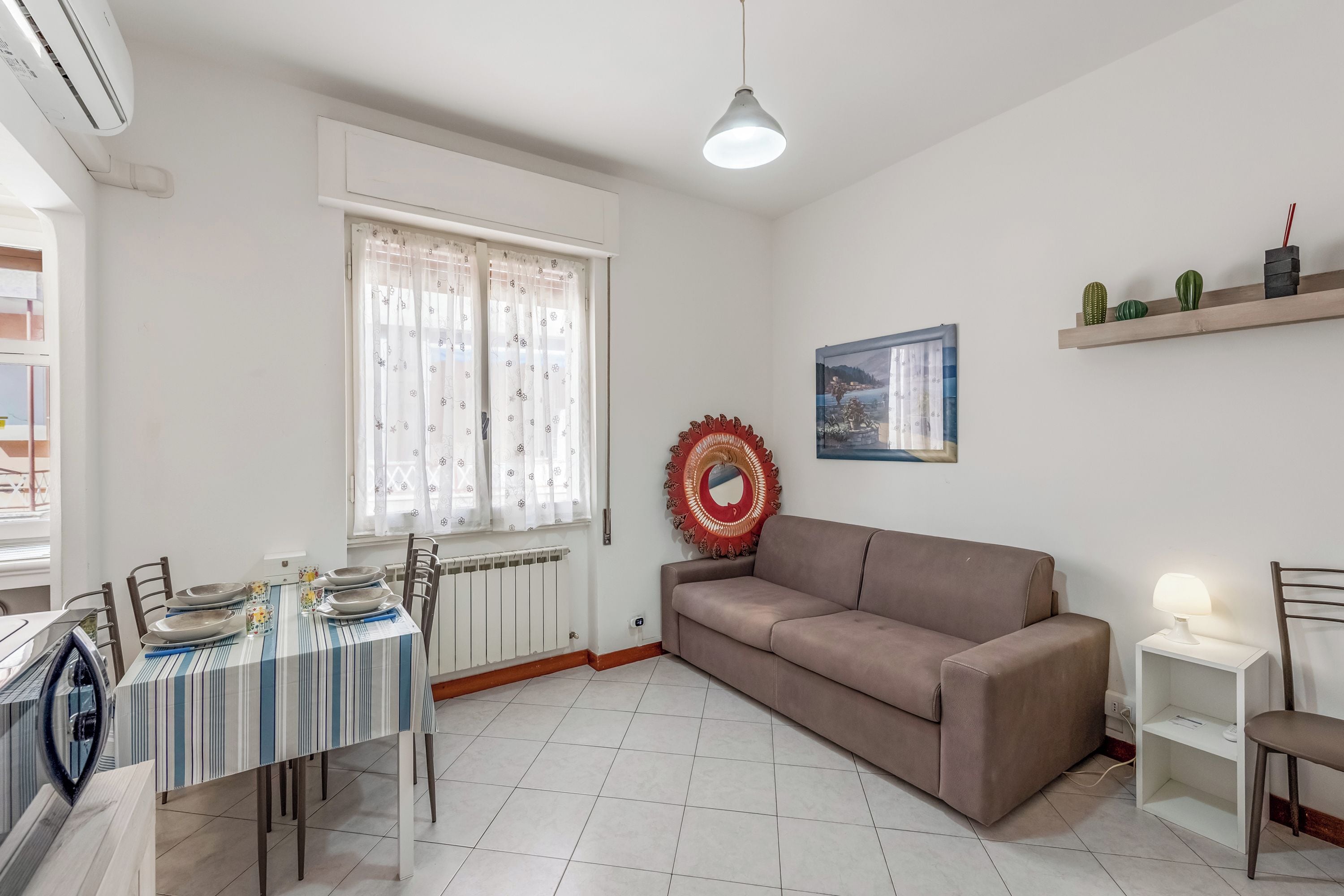 Sala de estar com sofá, mesa de jantar posta com pratos, radiador, janela com cortinas, prateleira com plantas e uma obra de arte na parede.