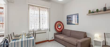 A living room with a sofa, a dining table set with plates, a radiator, a window with curtains, a wall-mounted shelf with plants, and a wall art piece.