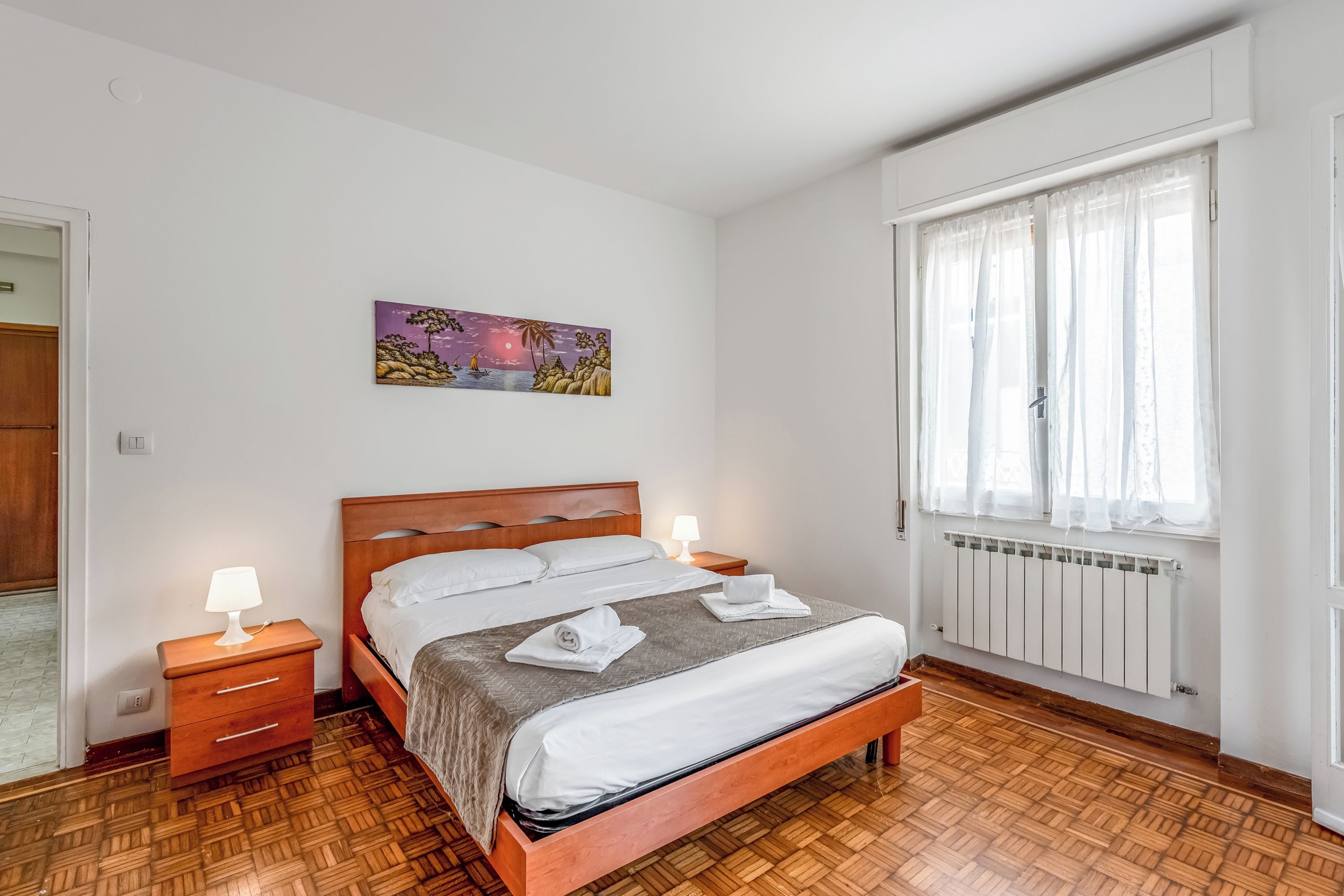 Um quarto com cama de madeira, roupa de cama branca, mesa de cabeceira com abajur, um quadro na parede e uma janela com cortinas finas.