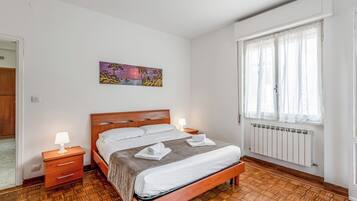 Um quarto com cama de madeira, roupa de cama branca, mesa de cabeceira com abajur, um quadro na parede e uma janela com cortinas finas.