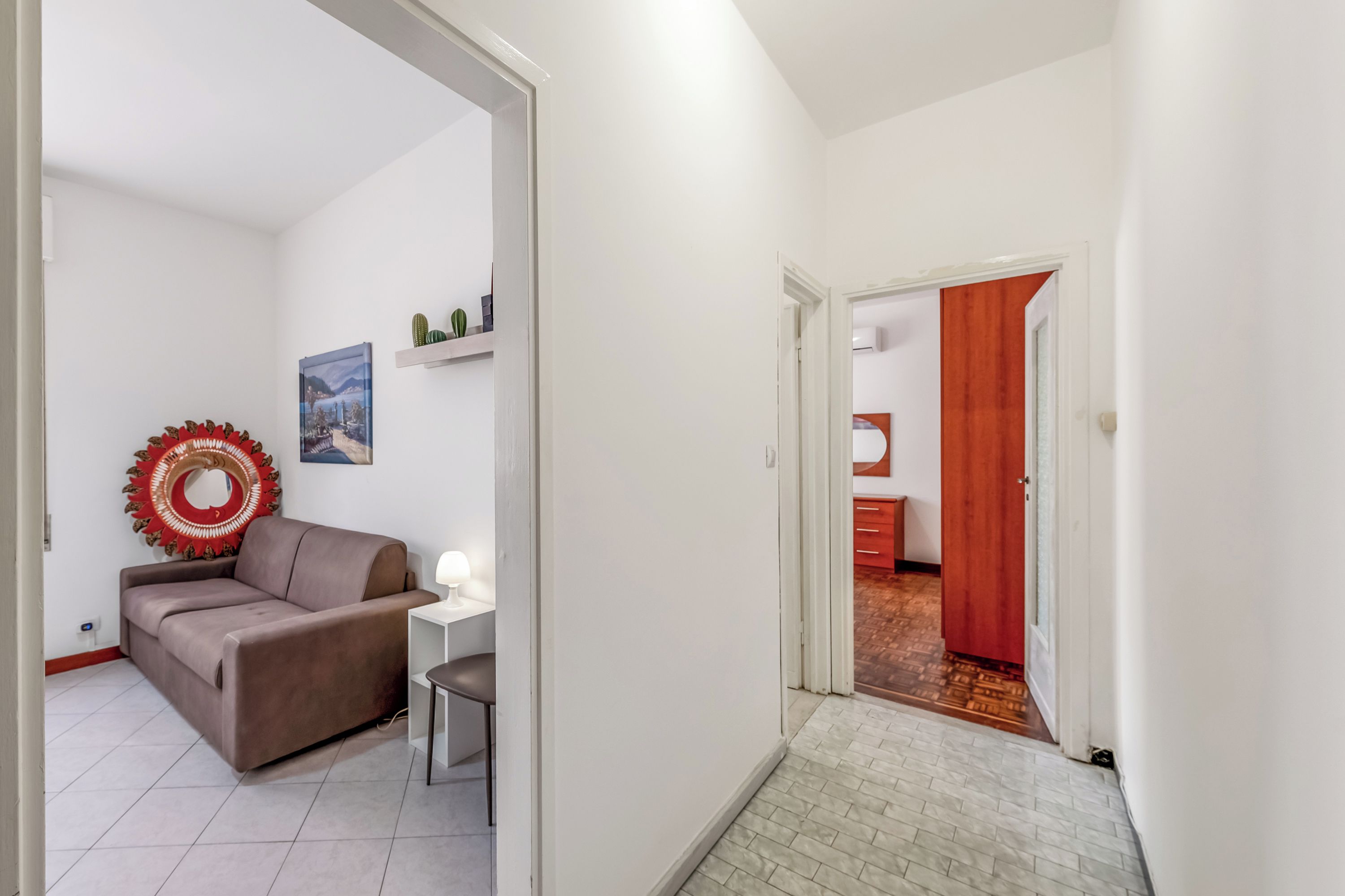 Uma sala de estar com um sofá, uma mesinha e uma luminária. Há uma porta que dá para outro quarto, com parede vermelha e piso de madeira.