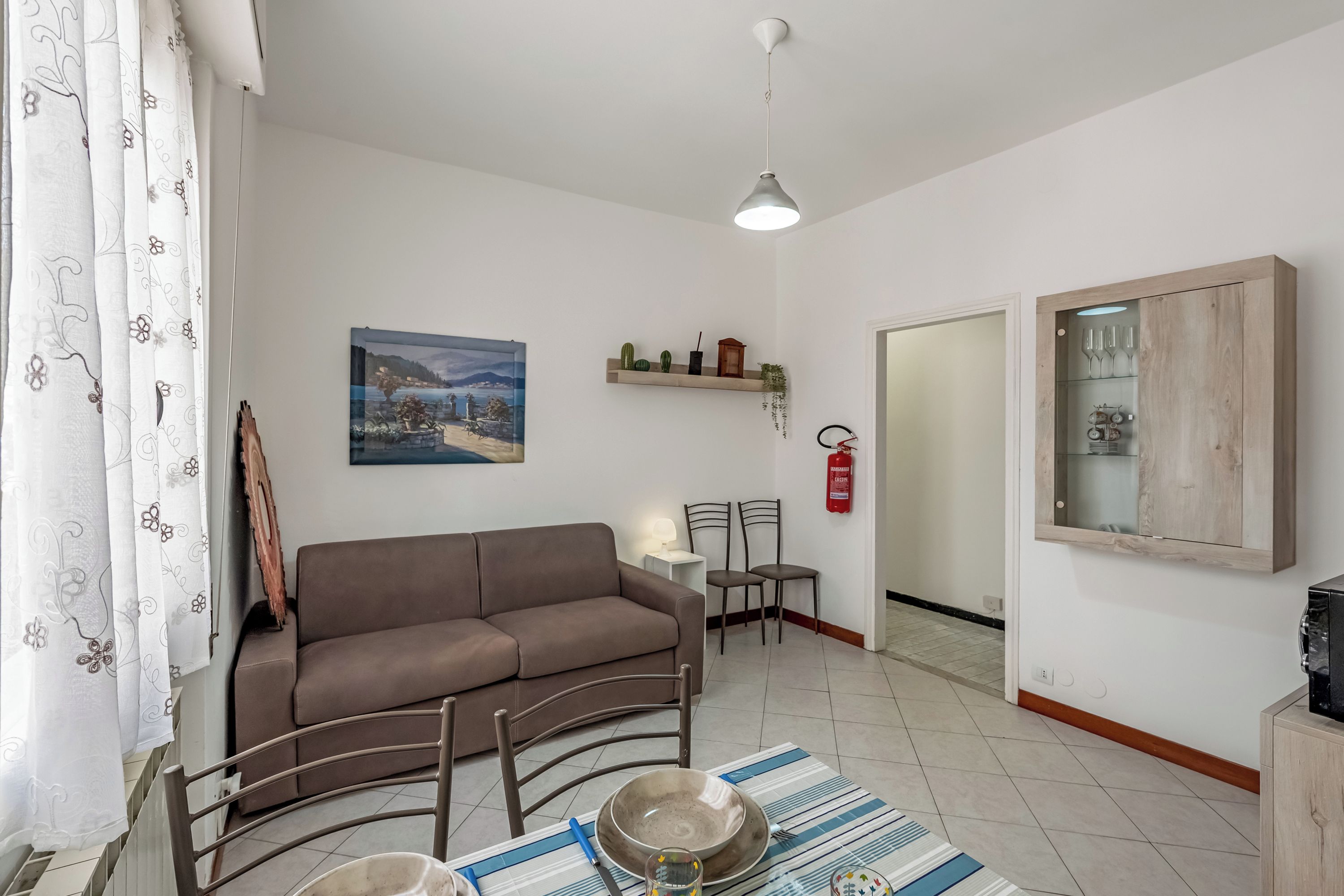 Uma sala de estar com um sofá marrom, mesa de jantar com cadeiras, um quadro na parede, uma estante com plantas e um armário com copos.