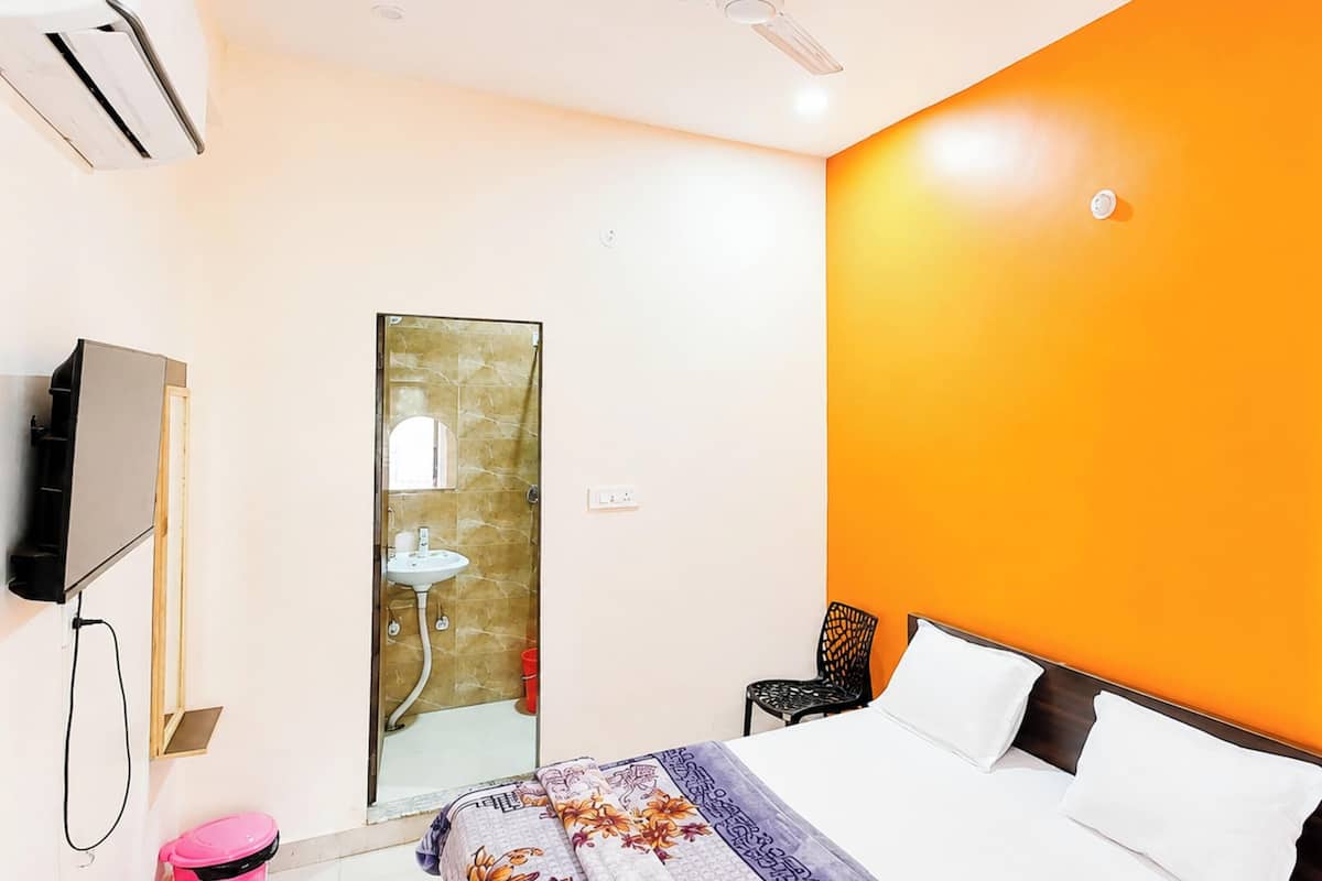 Habitación de hotel con cama, televisor montado en la pared, baño con lavamanos y espejo, y pared naranja.
