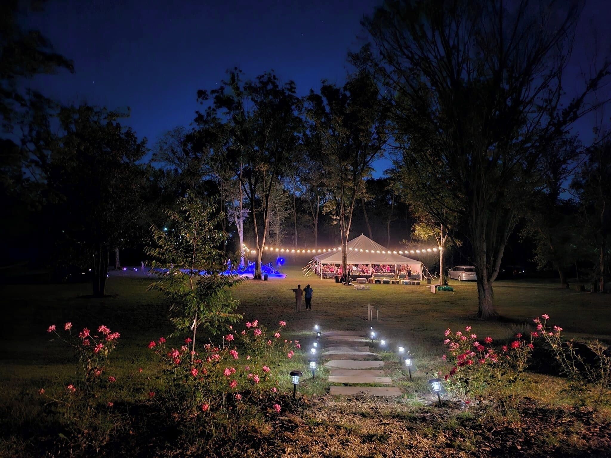 A nighttime outdoor event with a tent, illuminated by string lights, surrounded by trees and a garden with pink flowers.