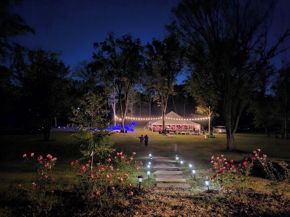 A nighttime outdoor event with a tent, illuminated by string lights, surrounded by trees and a garden with pink flowers.