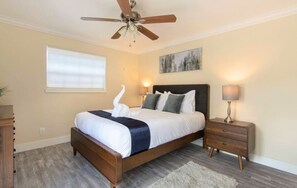 Ein Schlafzimmer mit einem Bett, Deckenventilator, Nachttisch, Lampe, einem Gemälde und einem Fenster mit Jalousien.