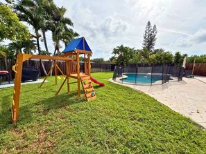 Ein Garten mit einer Holzspielanlage, einem Pool und einem eingezäunten Bereich.