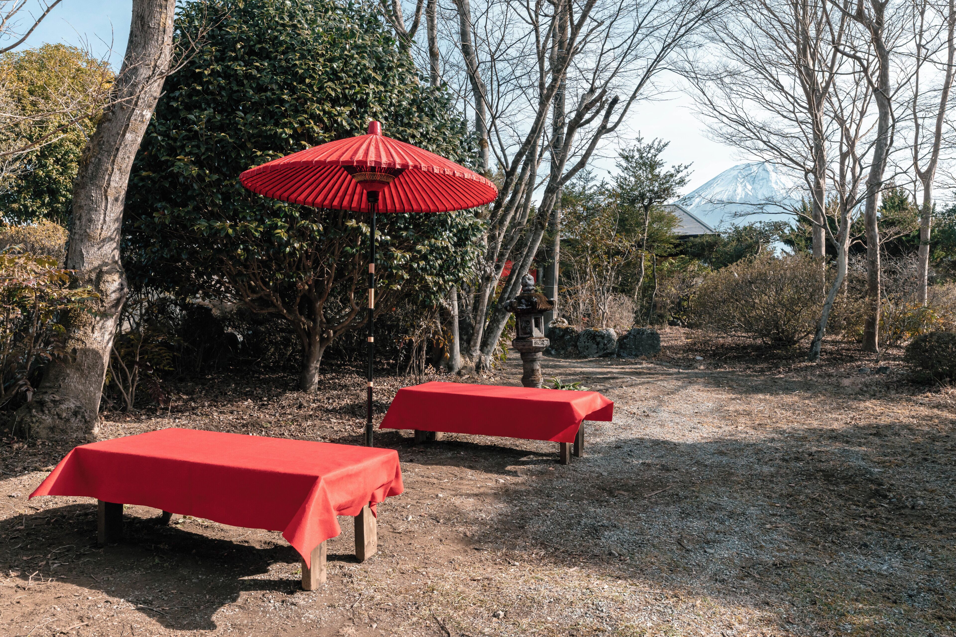 Two outdoor tables with red tablecloths, a red umbrella, and a stone lantern in a garden setting with trees and a mountain in the background.