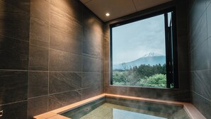 A modern bathroom with dark tiled walls, a glass-enclosed hot tub, and a view of a mountain. - nakafuji (Fujinomiya)