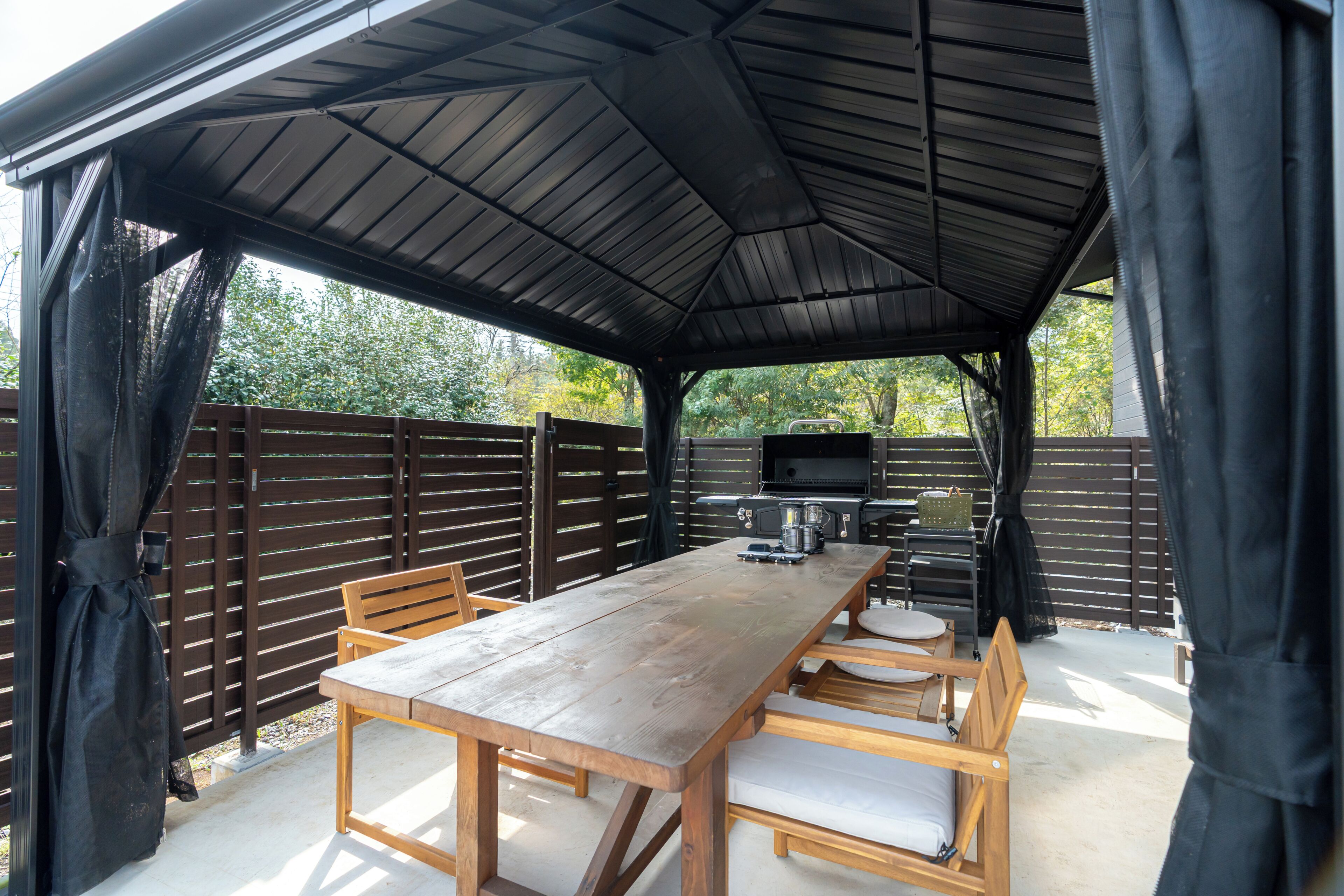 A covered outdoor dining area with a table and chairs, a grill, and a view of trees.