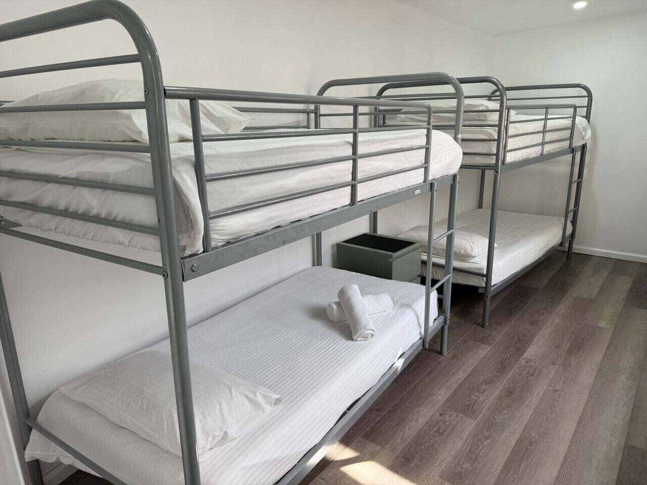 Dormitorio con literas de metal, ropa de cama blanca y una mesita de noche pequeña verde.