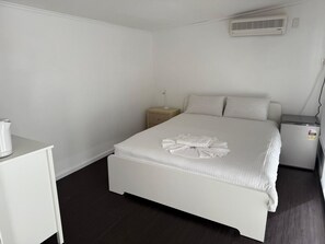 A white bed with a white pillow and a white blanket, a bedside table, a nightstand, a small heater, and a wall-mounted air conditioner.