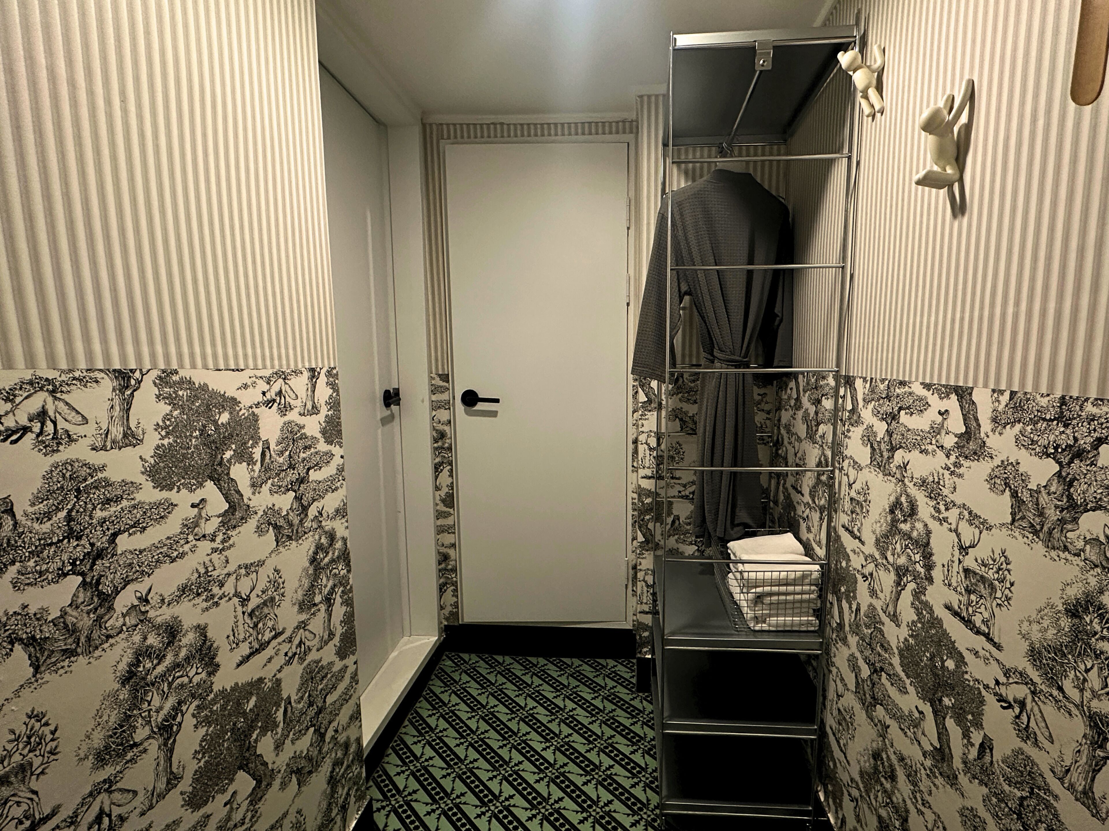 A narrow hallway with patterned wallpaper, a door, a metal shelf with towels, and a small mirror.