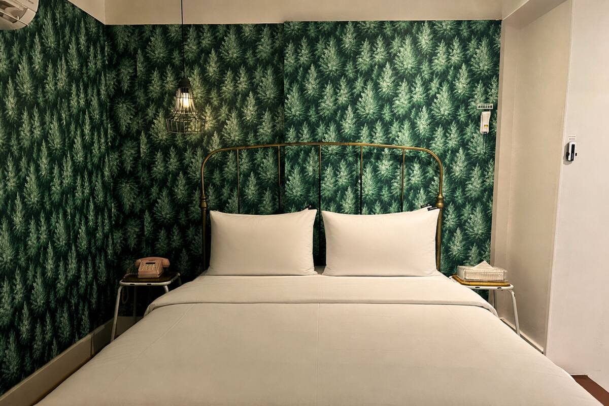 Una cama con estructura metálica y ropa de cama blanca, una mesita de noche con teléfono y una pared cubierta con papel tapiz de hojas verdes.