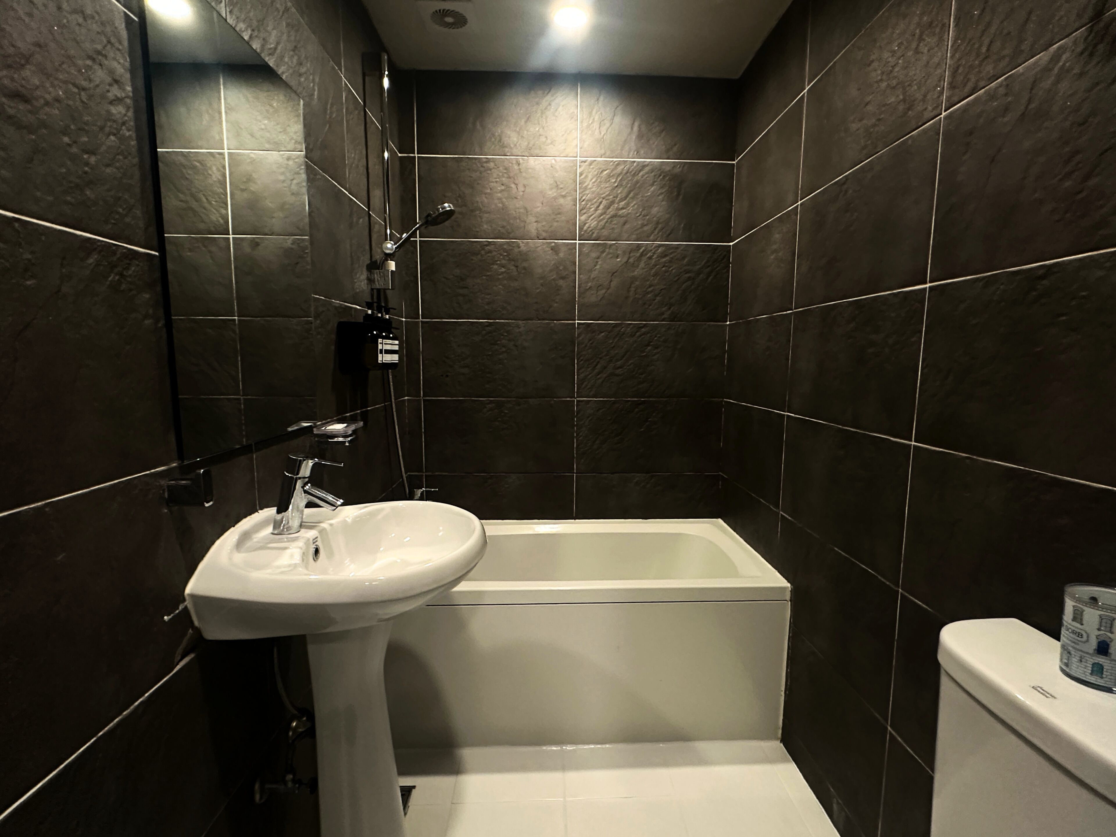 Un baño con paredes de baldosas oscuras, un lavamanos blanco y una bañera.