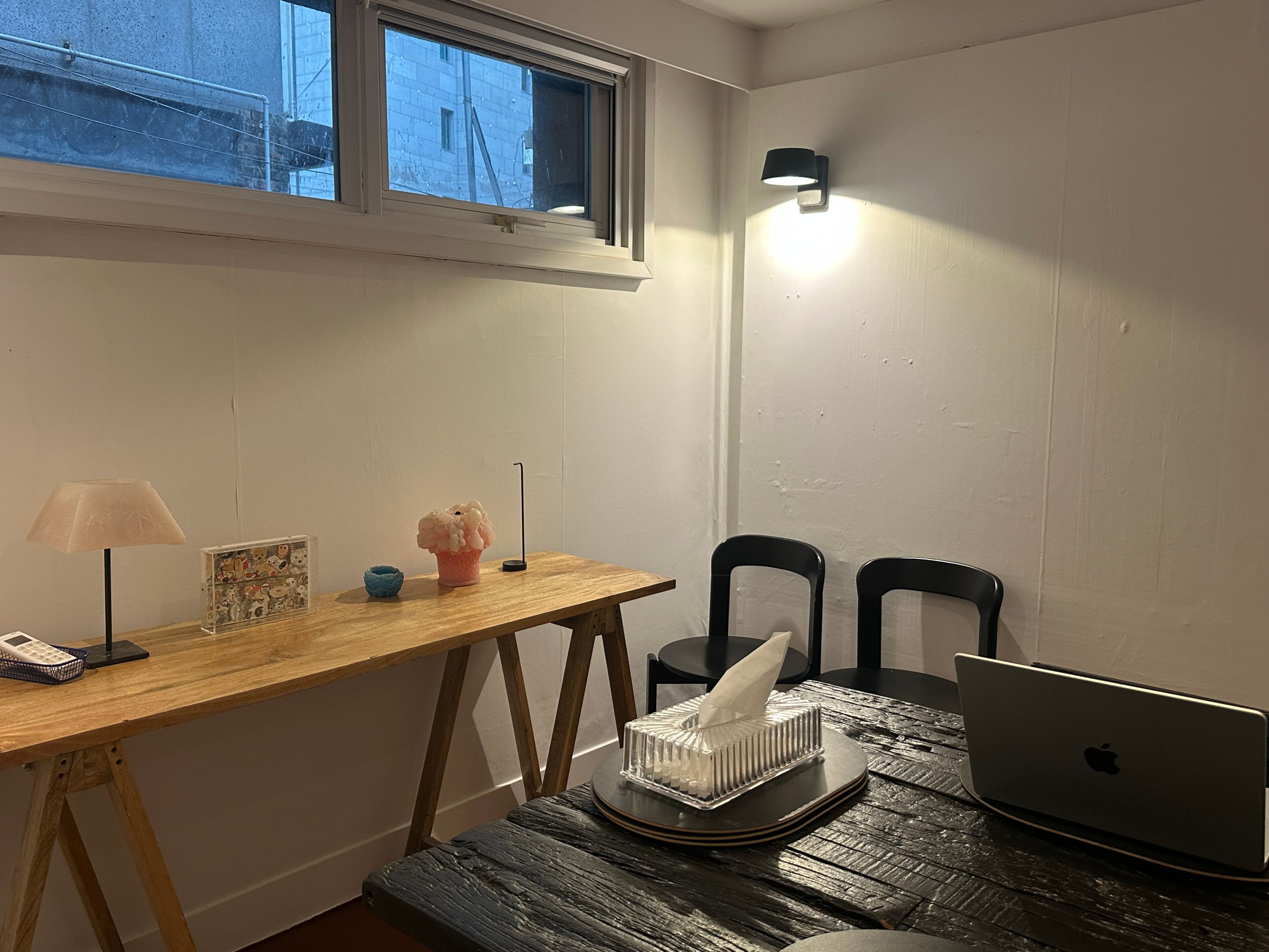 Une petite pièce avec un bureau en bois, un ordinateur portable, deux chaises, une lampe et une fenêtre.