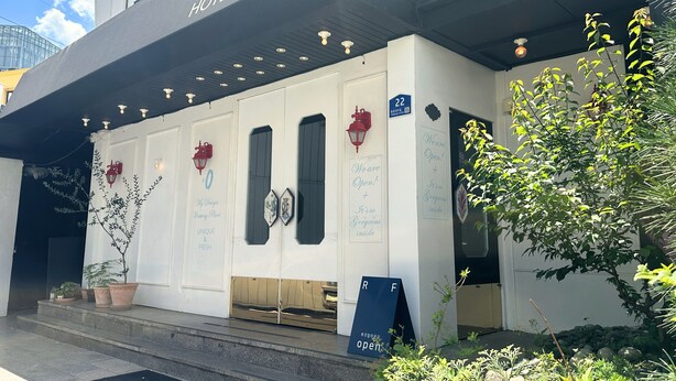 Een hotelentree met een bord waarop 'HOTEL LE O' staat, een rode lantaarn en een bord met de tekst 'Geopend' en 'Het is er zo prachtig van binnen'.