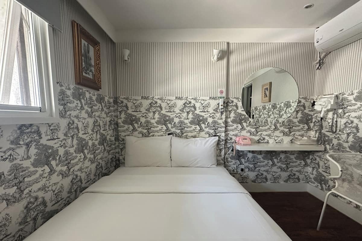 Una habitación con una cama, un espejo, un estante con teléfono y cepillo, y papel tapiz estampado.