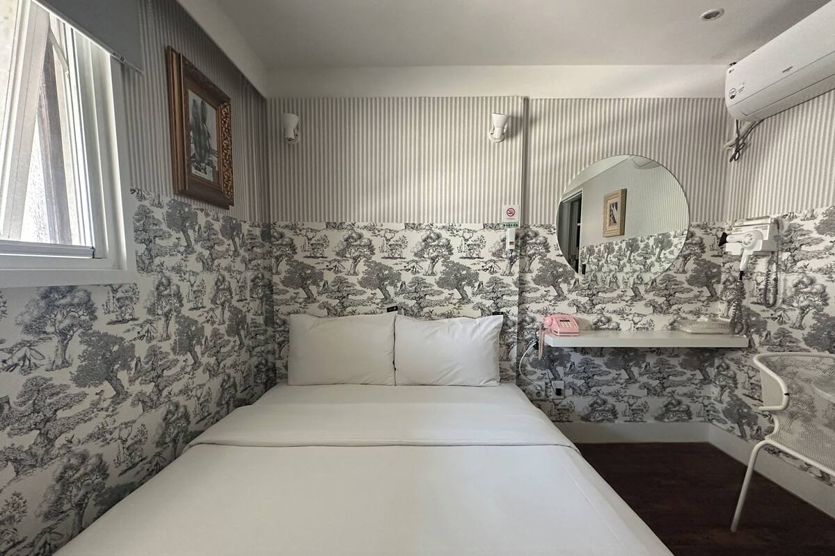 Een kamer met een bed, een spiegel, een plankje met een telefoon en een borstel, en behang met een patroon.