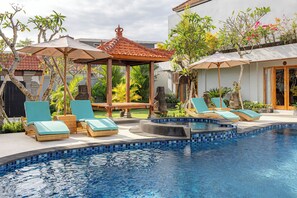Ein Swimmingpool mit Liegestühlen und Sonnenschirmen, umgeben von tropischen Pflanzen und einem Pavillon.