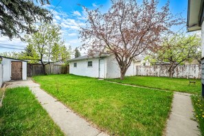 A residential backyard with a white house, a wooden fence, a tree, and a paved path. - 3BR bungalow near Chinook Mall, office, sleeps 8 (Calgary)