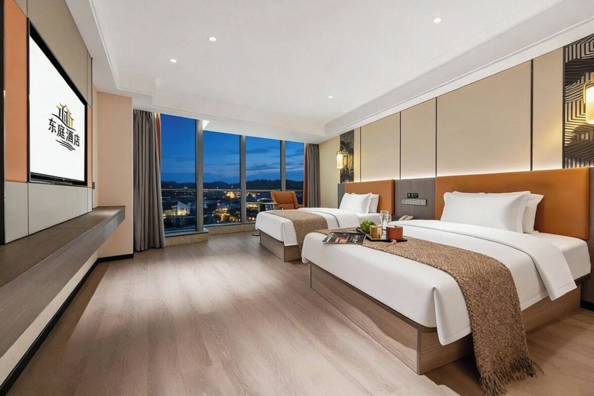 Ein Hotelzimmer mit zwei Betten, einem großen Fenster mit Blick auf die Stadt, einem Fernseher und einem Holzboden.