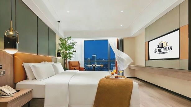A modern hotel room with a bed, bedside table, lamp, chair, and a city view through a large window.
