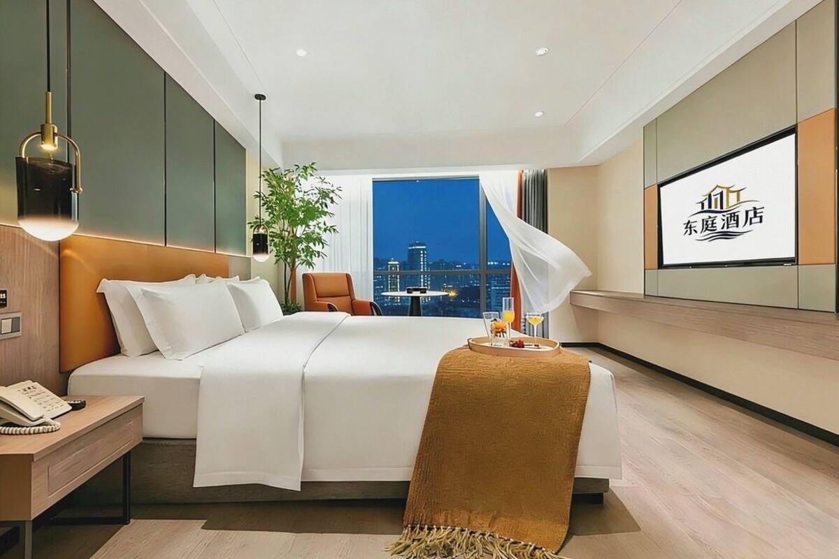 Ein modernes Hotelzimmer mit Bett, Nachttisch, Lampe, Sessel und Blick auf die Stadt durch ein großes Fenster.