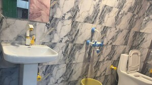 A bathroom with a white sink, a toilet, and a yellow bucket. - Hotel Youvraj Inn (Ghaziabad)