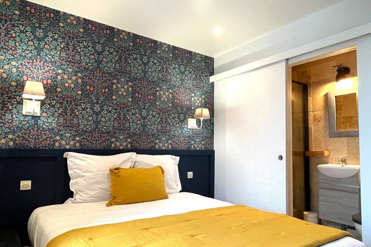 A hotel room with a bed, a nightstand, a wall with floral wallpaper, two wall-mounted lamps, and a bathroom with a sink and mirror.