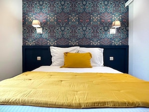 Uma cama com colcha amarela e almofada cor mostarda, cabeceira azul escura e papel de parede estampado com flores.
