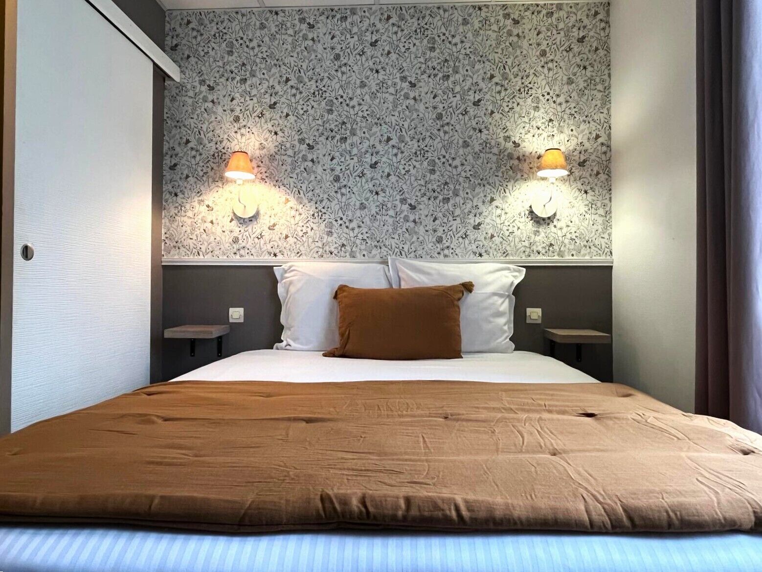 Habitación de hotel con cama, dos lámparas fijadas a la pared, papel tapiz estampado y un cojín marrón.