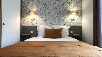 Habitación de hotel con cama, dos lámparas fijadas a la pared, papel tapiz estampado y un cojín marrón.