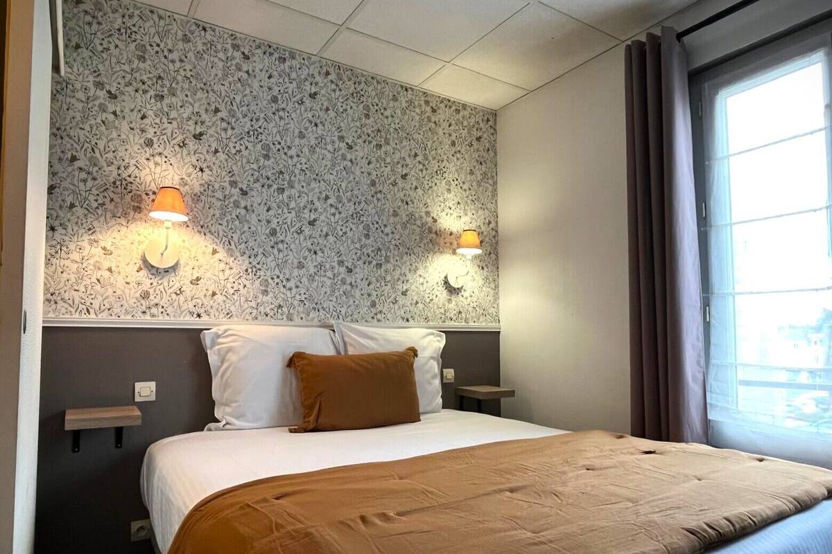 A hotel room with a bed, two wall-mounted lamps, a window with curtains, and a floral-patterned wall.