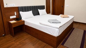 A hotel room with a wooden headboard, a single bed with white bedding, a nightstand with a telephone, and a wooden door. - Ram Sadan Ayodhya (Ayodhya)