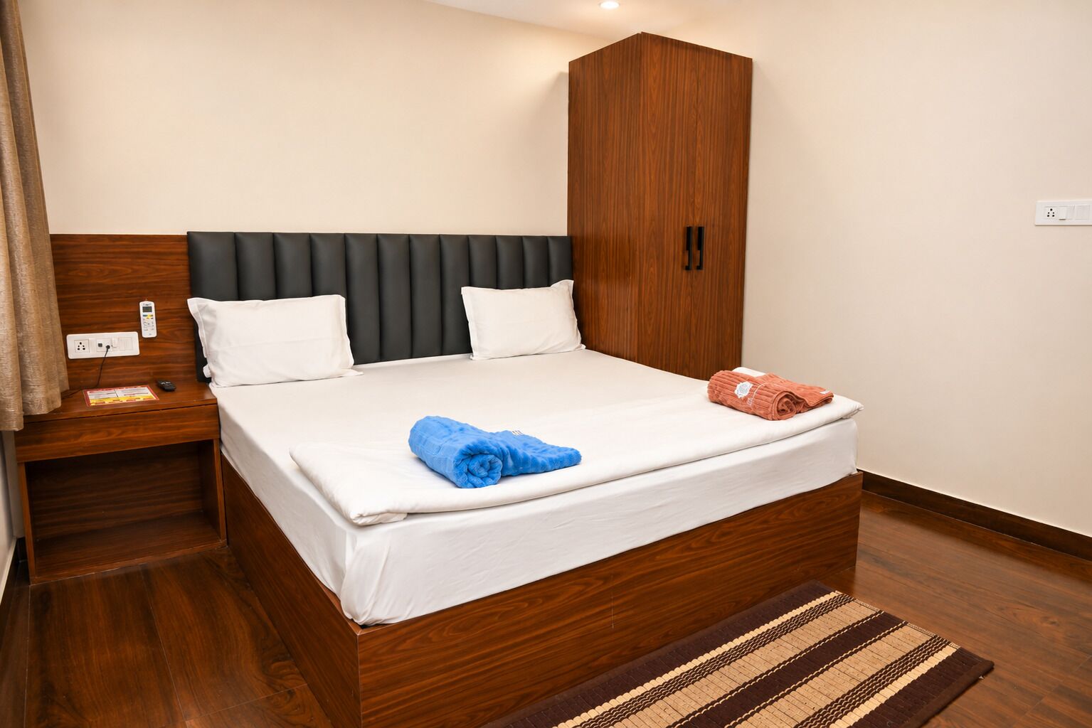 A hotel room with a wooden bed frame, white bedding, a dark headboard, a wooden wardrobe, and a striped rug.