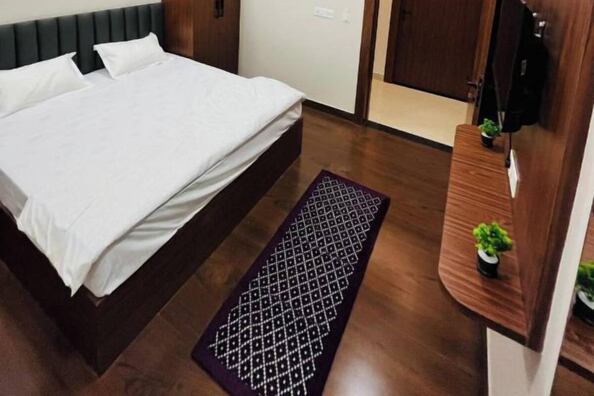 A bedroom with a bed, a wooden wardrobe, a television mounted on the wall, and a door leading to another room.