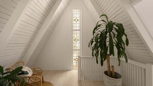 Quarto de sótão amplo, com teto alto, uma grande janela, um vaso de planta e uma cadeira de vime.