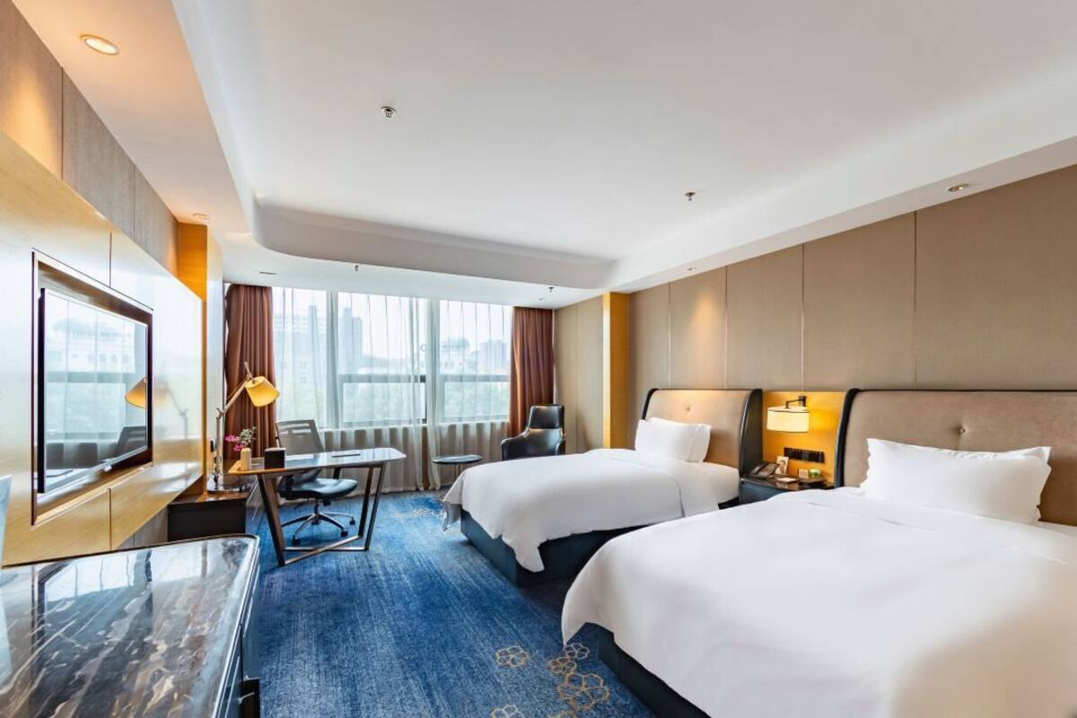 A hotel room with two beds, a desk, a TV, and a large window.